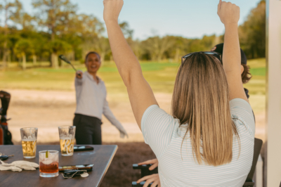 A friend cheers from a table of drinks as a golfer celebrates a shot on the course