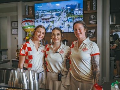 Three Pine Royale servers in checkered uniforms smiling behind the bar
