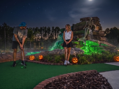 A couple playing a Halloween-lit hole of Mini Royale at night, jack-o'-lanterns along the green and a skeleton on the waterfall