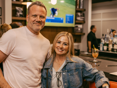 A smiling couple at the Pine Royale bar, a cocktail on the counter and golf on the TV behind
