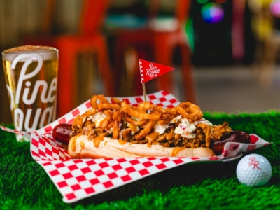A specialty hot dog piled with pulled pork, slaw and crispy onions, beside a beer in a Pine Royale glass