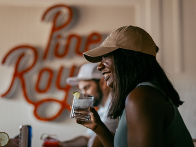 A woman in a cap laughing with a drink in hand beneath the Pine Royale sign