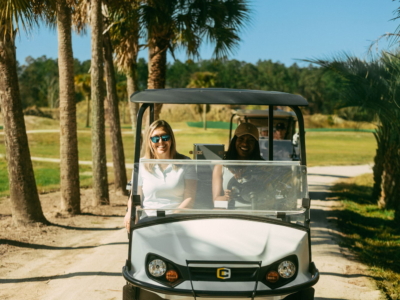 Two women driving a golf cart along a palm-lined cart path