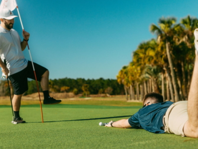 A golfer lying flat on the green to line up a putt as a friend holds the flag