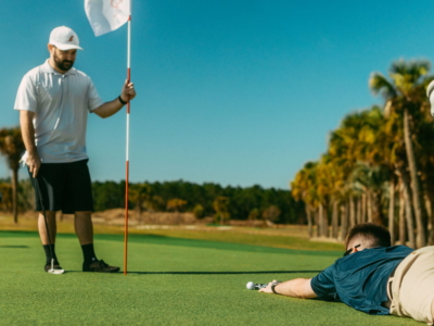 A golfer stretched out on the green reading his putt while a friend tends the pin