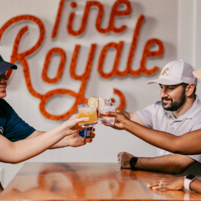 Four friends clinking drinks beneath the lit Pine Royale sign in the restaurant
