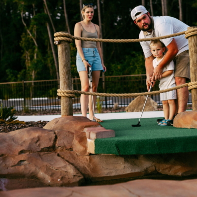 A father helping his young son putt on the rope-bridge hole at Mini Royale as mom watches