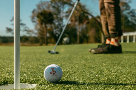 A PR-monogrammed ball beside the cup as a golfer in checkered socks putts