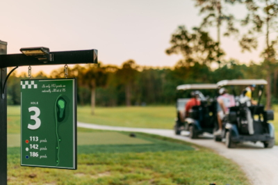 The hole 3 sign — 113, 142 and 186 yards, and "It's only 140 yards so naturally I'll hit it 95" — with golf carts passing behind