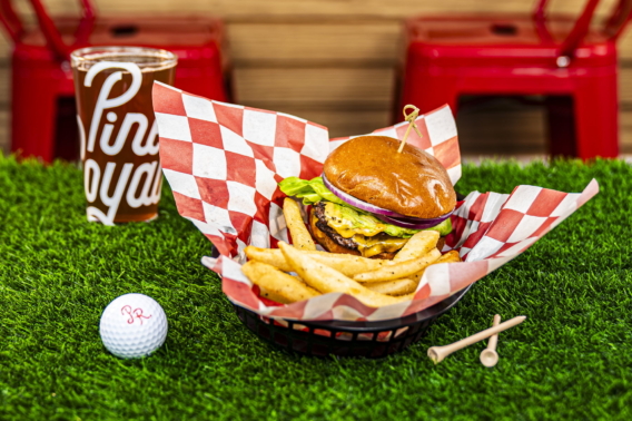 The Hole-In-One Burger with fries in a checkered basket, beside a Pine Royale glass