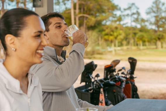 A golfer sipping a drink beside a smiling friend, golf bags lined up behind them