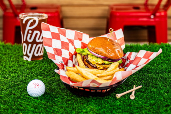 The Hole-In-One Burger and fries in a checkered basket with a Pine Royale glass and golf ball