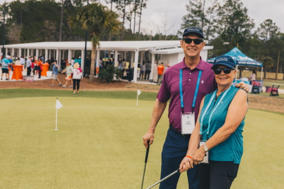 A couple in event lanyards posing with putters on the practice green during an outing