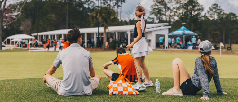 A family sitting on the grass watching an event, a picnic bag beside them