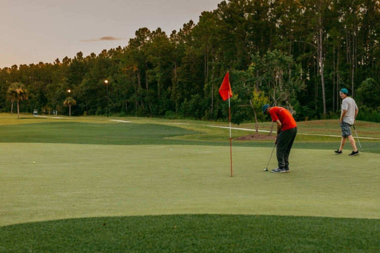 A golfer in red putting toward the flag at sunset as a friend walks off the green