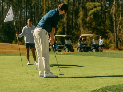 A golfer putting while a friend tends the flag, golf carts parked behind the green